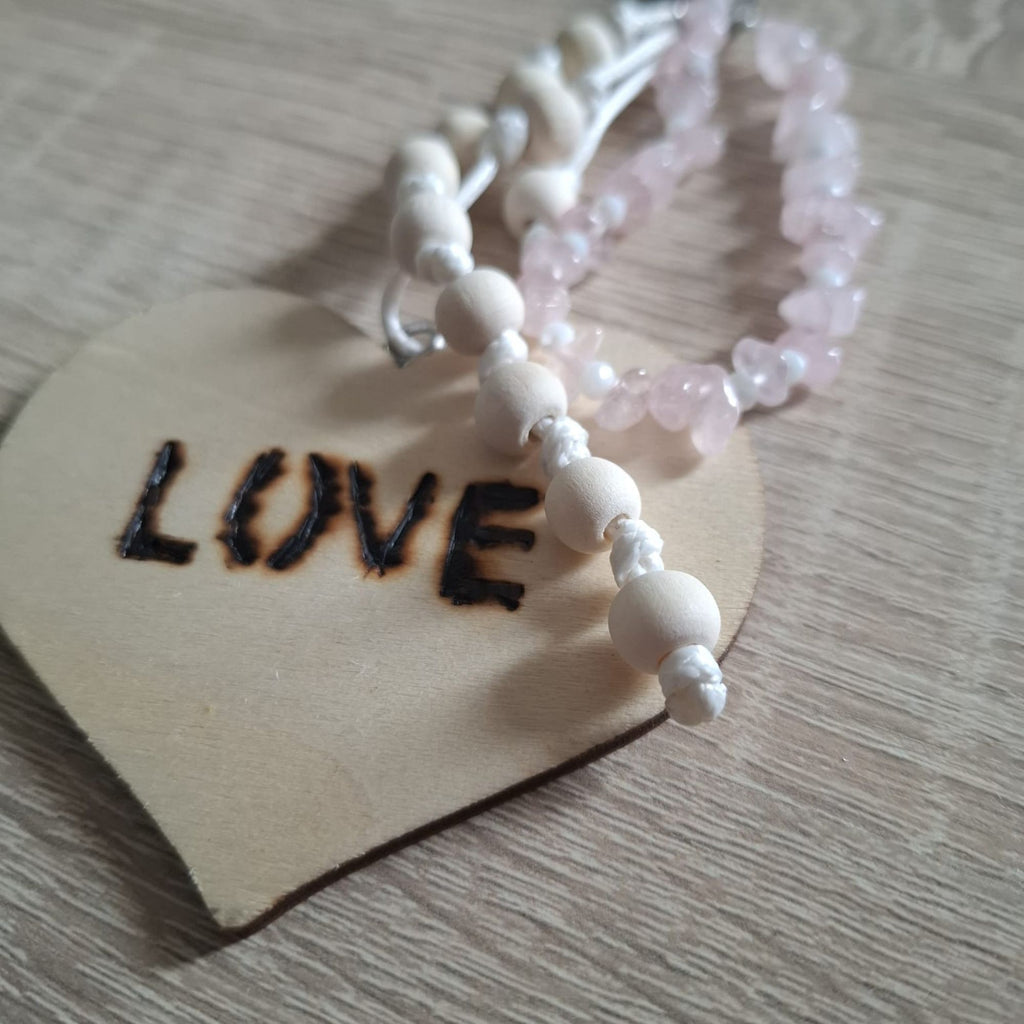 Handmade Amora bag charm featuring genuine pale pink rose quartz chip beads, small white accent beads and natural wooden beads hand-knotted on white cord. A large natural wooden heart with the burned-in word “LOVE” creates the romantic centerpiece, while a silver-tone heart-shaped clasp completes this unique boho-inspired gemstone handbag accessory.
