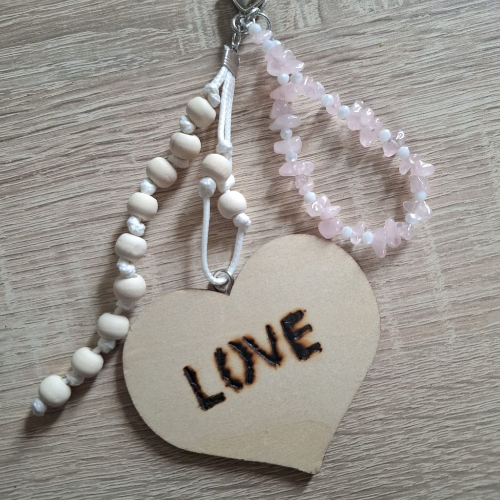 Close-up detail of the handmade Amora bag charm, showing the large natural wooden LOVE heart, hand-knotted white cord, smooth wooden beads and delicate pale pink rose quartz gemstone chips with small white accent beads. The close-up highlights the natural textures, soft romantic colors and handcrafted details of this unique boho bag accessory.