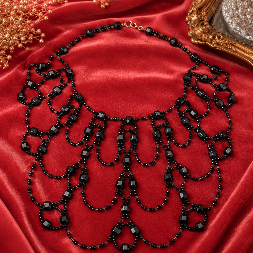 Black Royal handgefertigte Art-Déco Statement-Halskette aus schwarzen und klaren Glasperlen, dekorativ auf rotem Satin präsentiert. Die aufwendig gearbeitete Halskette besteht aus zahlreichen symmetrisch angeordneten Perlenschlaufen mit kleinen schwarzen Glasperlen und facettierten schwarzen Würfelperlen. Der breite, mehrreihige Aufbau verleiht dem Collier eine elegante und außergewöhnliche Optik für Abendmode und festliche Anlässe.