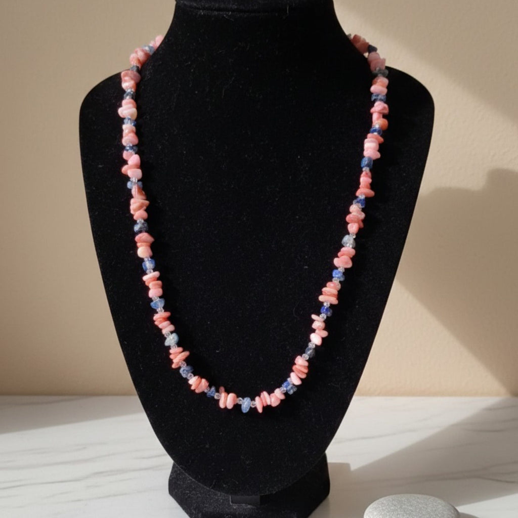 Coral Blossom handmade gemstone necklace displayed on a black jewelry bust in warm sunlight. Irregular pink coral chips alternate with small blue apatite stones and clear sparkling accents, creating a delicate pink-and-blue color contrast. The dark velvet display highlights the natural shapes and individual variations of the stones.