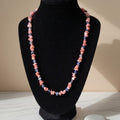 Coral Blossom handmade gemstone necklace displayed on a black jewelry bust in warm sunlight. Irregular pink coral chips alternate with small blue apatite stones and clear sparkling accents, creating a delicate pink-and-blue color contrast. The dark velvet display highlights the natural shapes and individual variations of the stones.