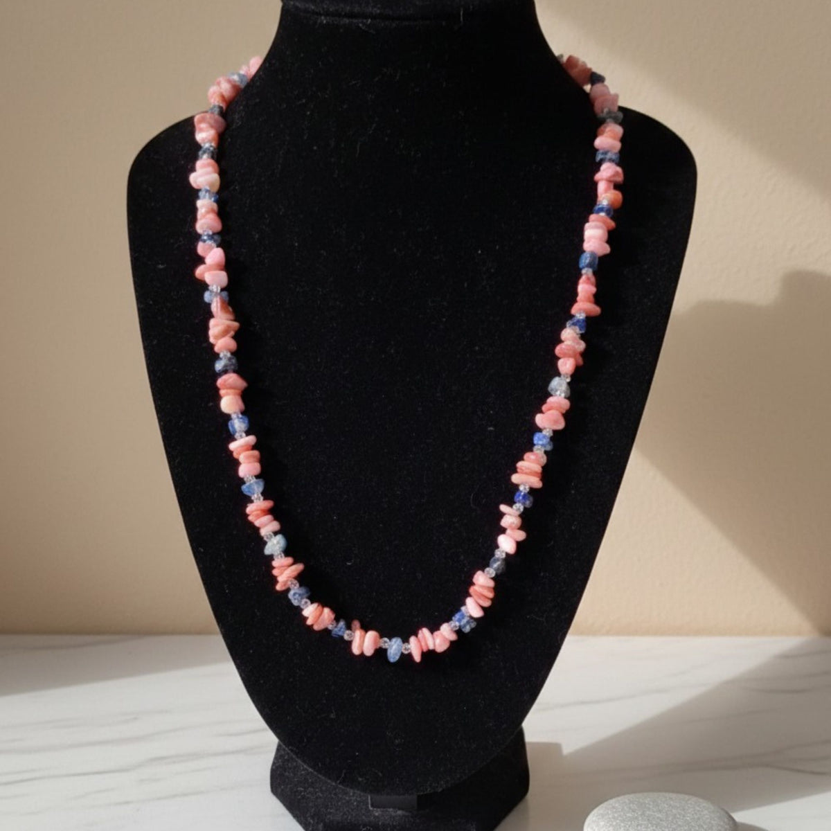 Coral Blossom handmade gemstone necklace displayed on a black jewelry bust in warm sunlight. Irregular pink coral chips alternate with small blue apatite stones and clear sparkling accents, creating a delicate pink-and-blue color contrast. The dark velvet display highlights the natural shapes and individual variations of the stones.