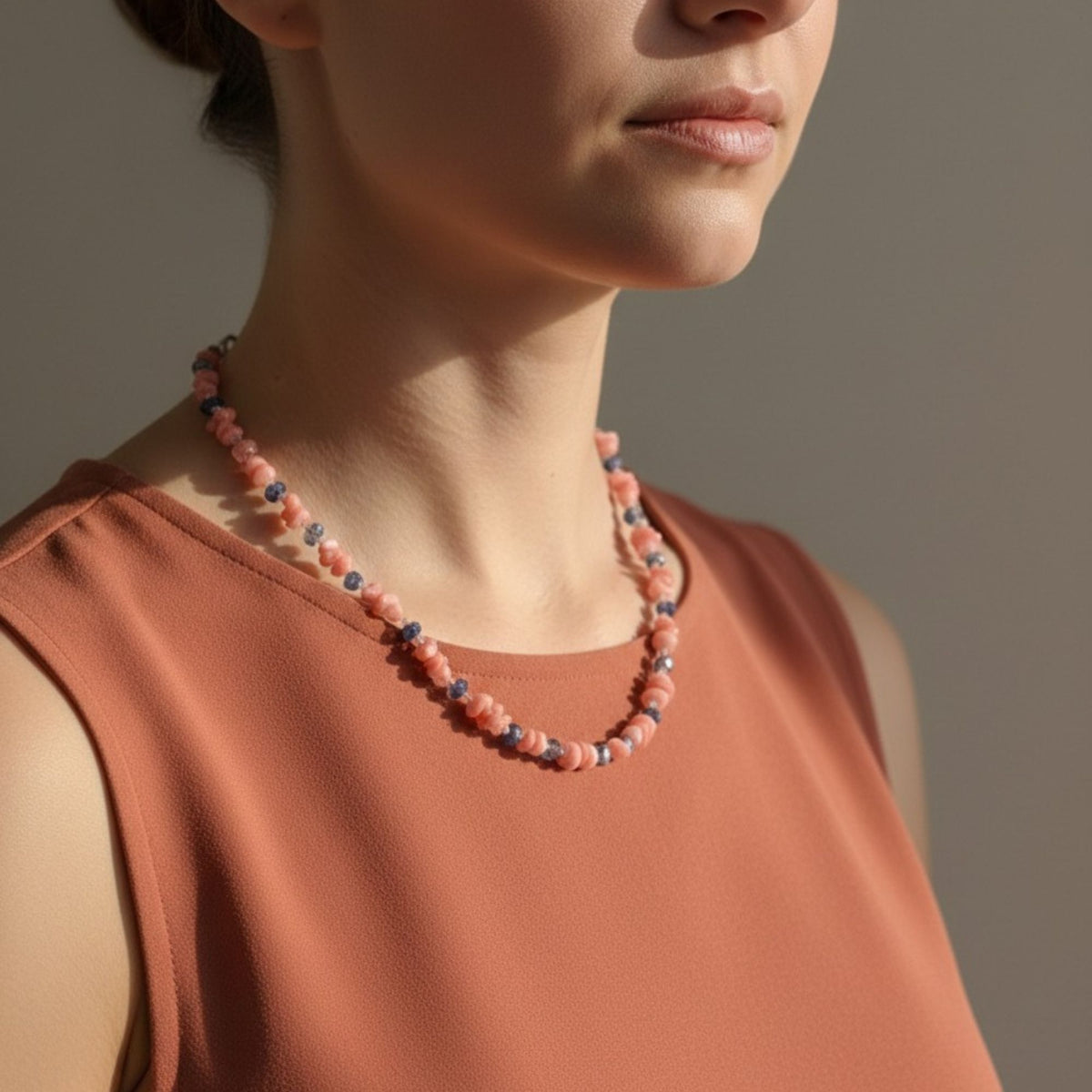Coral Blossom handmade Pink Coral and Blue Apatite necklace worn by a woman with a warm terracotta sleeveless top. The irregular pink coral chips are accented by small blue apatite stones and clear sparkling beads, showing the necklace’s natural texture, color contrast and approximately 50 cm length when worn.
