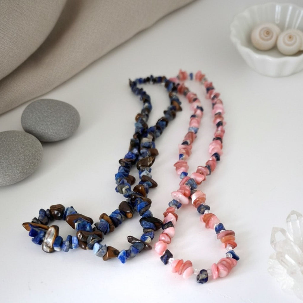 Coral Blossom and Warrior’s Sky gemstone necklaces displayed together on a white surface. Coral Blossom features pink coral and blue apatite, while Warrior’s Sky combines deep blue lapis lazuli with golden-brown tiger’s eye, presenting the two handmade necklaces as a symbolic partner set.