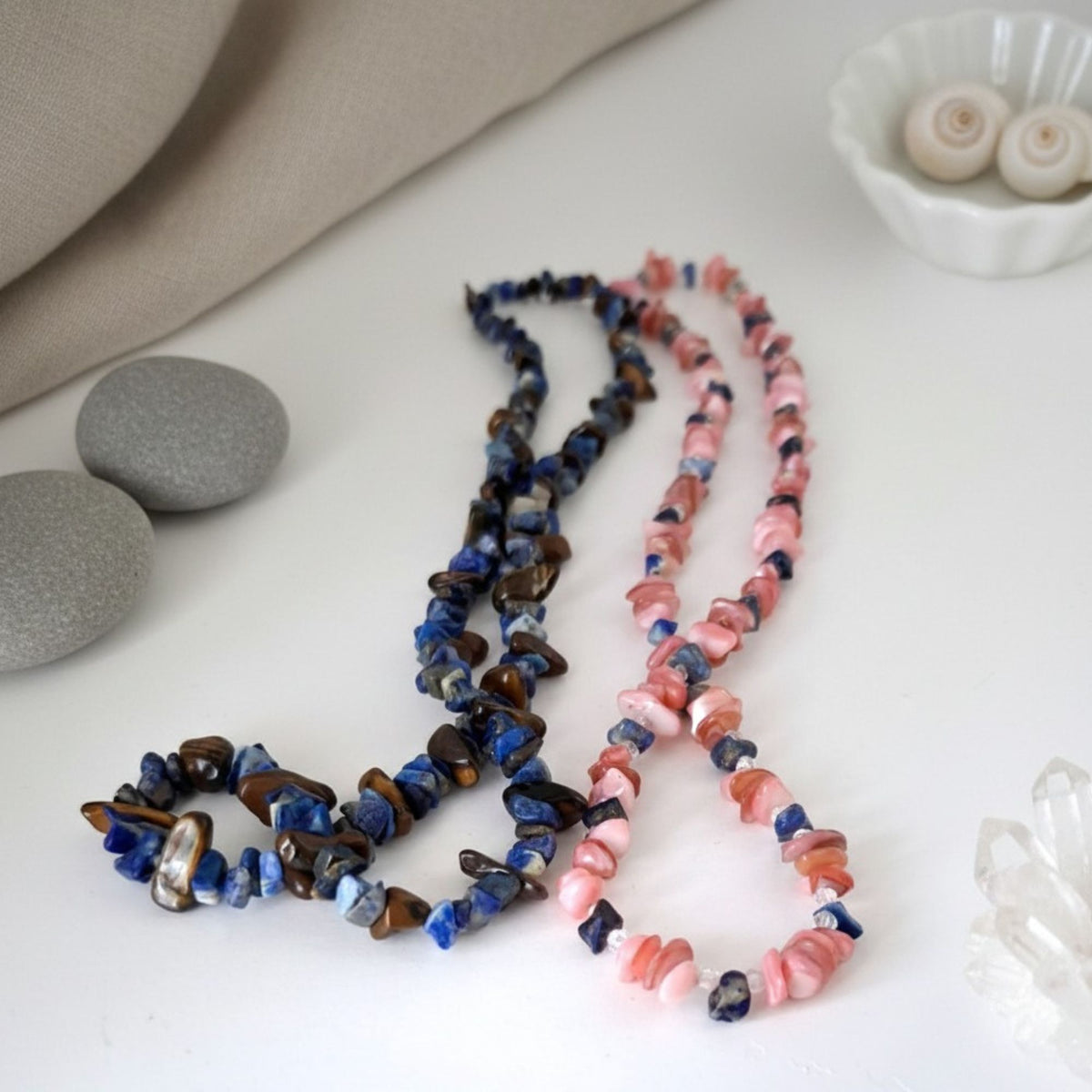 Coral Blossom and Warrior’s Sky gemstone necklaces displayed together on a white surface. Coral Blossom features pink coral and blue apatite, while Warrior’s Sky combines deep blue lapis lazuli with golden-brown tiger’s eye, presenting the two handmade necklaces as a symbolic partner set.