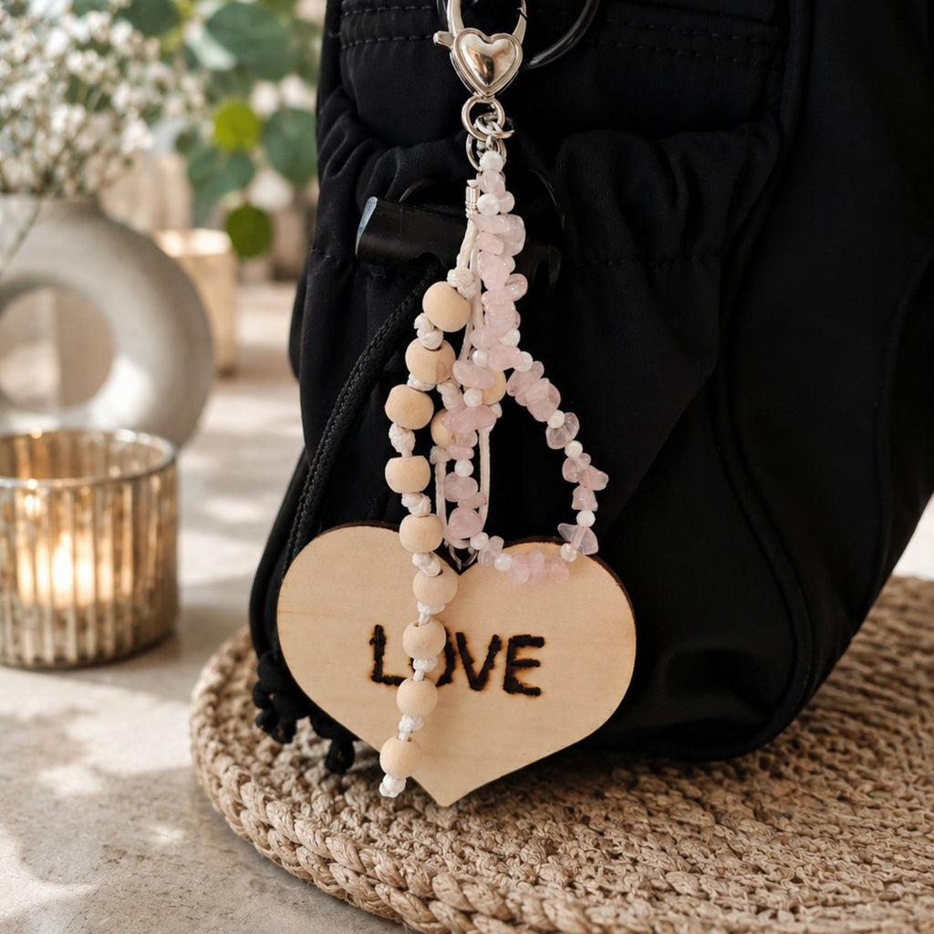 Handmade Amora bag charm with pale pink rose quartz gemstone chips, small white accent beads and natural wooden beads knotted on white cord. The statement accessory features a large wooden heart with burned-in “LOVE” lettering and a silver-tone heart-shaped clasp, shown hanging from a black handbag in a warm elegant setting.