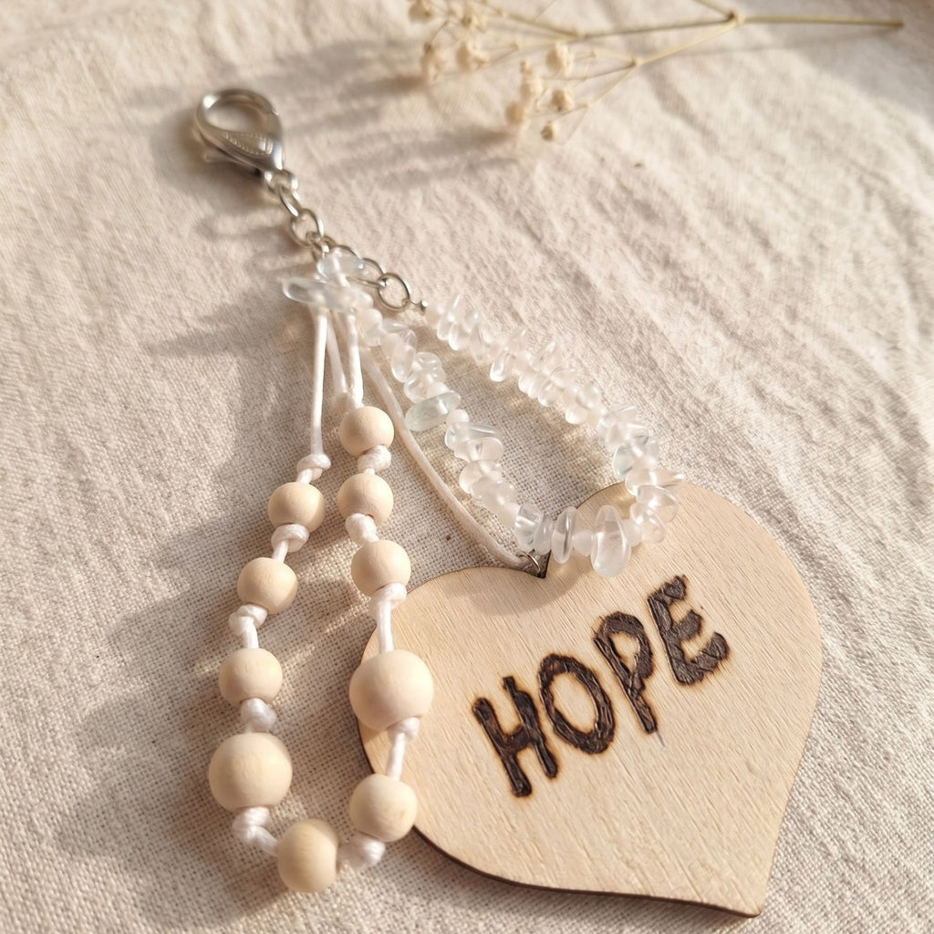 Detail view of the handmade Hope bag charm featuring genuine clear quartz chips, natural wooden beads on white knotted cords and a large wooden heart engraved with “HOPE”. The silver-tone lobster clasp is visible at the top, while warm natural light highlights the textures of the clear gemstones, unfinished wood and cord against soft linen fabric.