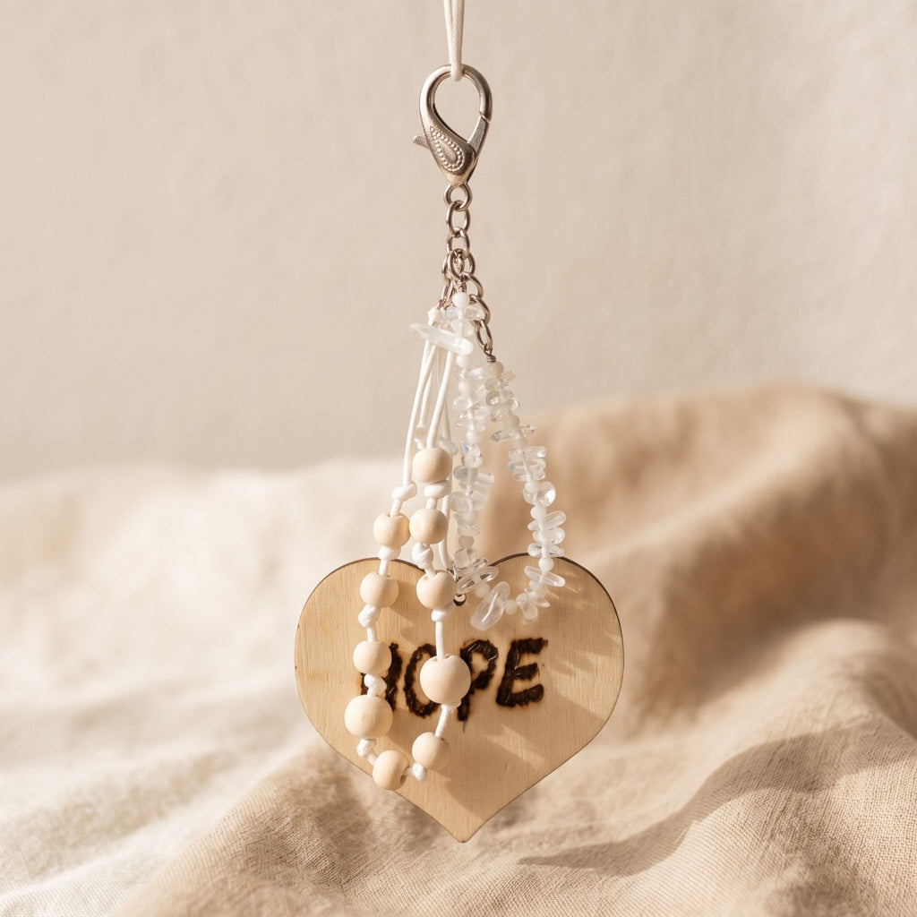 Handmade Hope bag charm displayed against soft beige fabric, featuring genuine clear quartz chips, natural wooden beads on white knotted cords and a large wooden heart engraved with “HOPE”. The full silver-tone lobster clasp and chain are visible, while warm sunlight highlights the transparent gemstones, natural wood and handcrafted details.