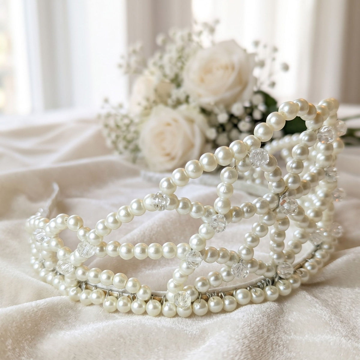 Close-up of the handmade Ivory Dream pearl bridal crown with ivory pearls and clear crystal beads, displayed on soft fabric with white roses in the background.