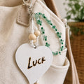 Handmade LUCK bag charm attached to a beige fabric handbag, featuring green aventurine chips, dark green and clear glass beads, natural wooden beads and white cord. A large white wooden heart with the hand-burned word “LUCK” completes the design, finished with a silver-colored lobster clasp.