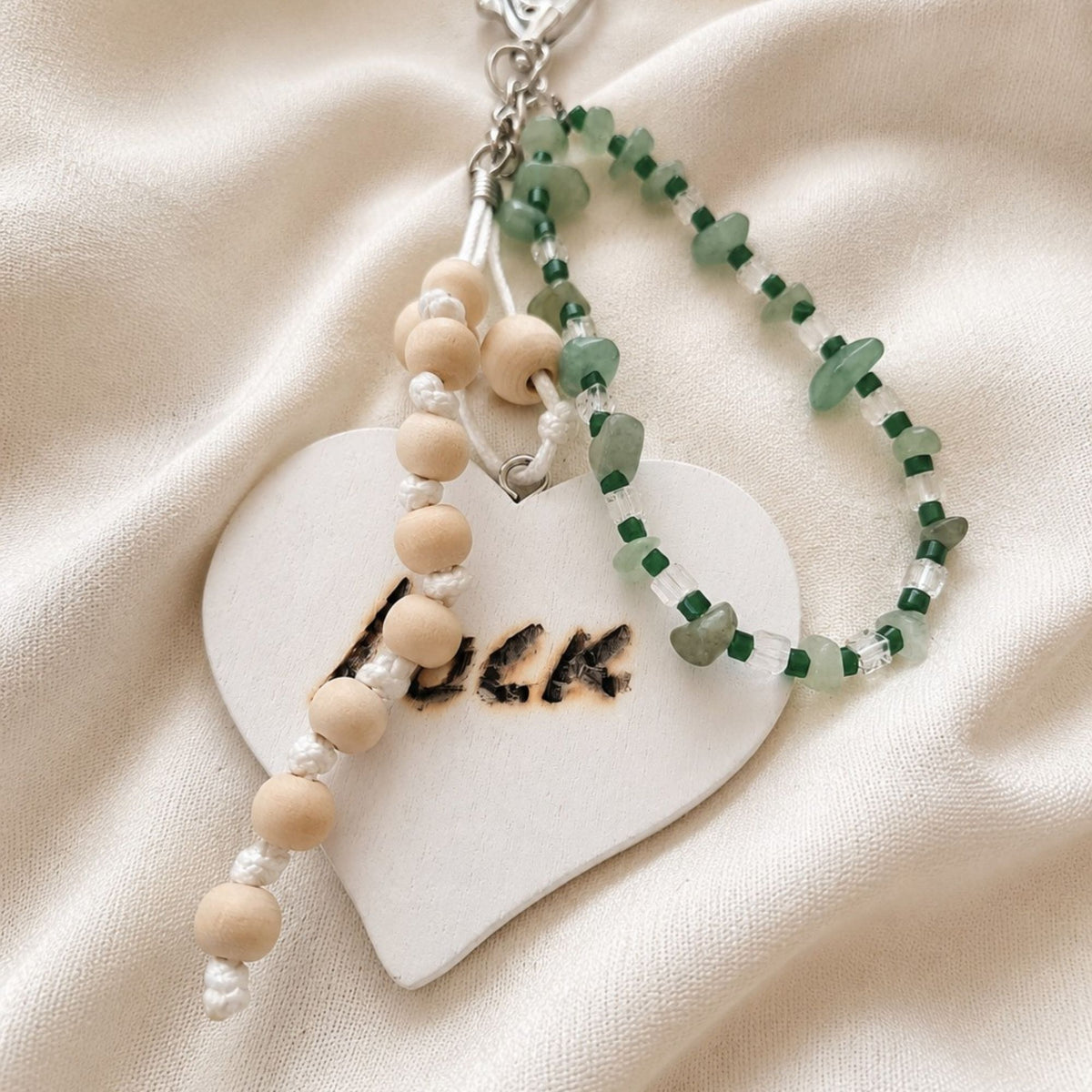 Handmade LUCK bag charm displayed on ivory satin, featuring green aventurine chips, dark green and clear glass beads, natural wooden beads and white cord. The wooden bead strand lies across the large white heart with the hand-burned word “LUCK”, showing the layered handmade design and silver-colored clasp.