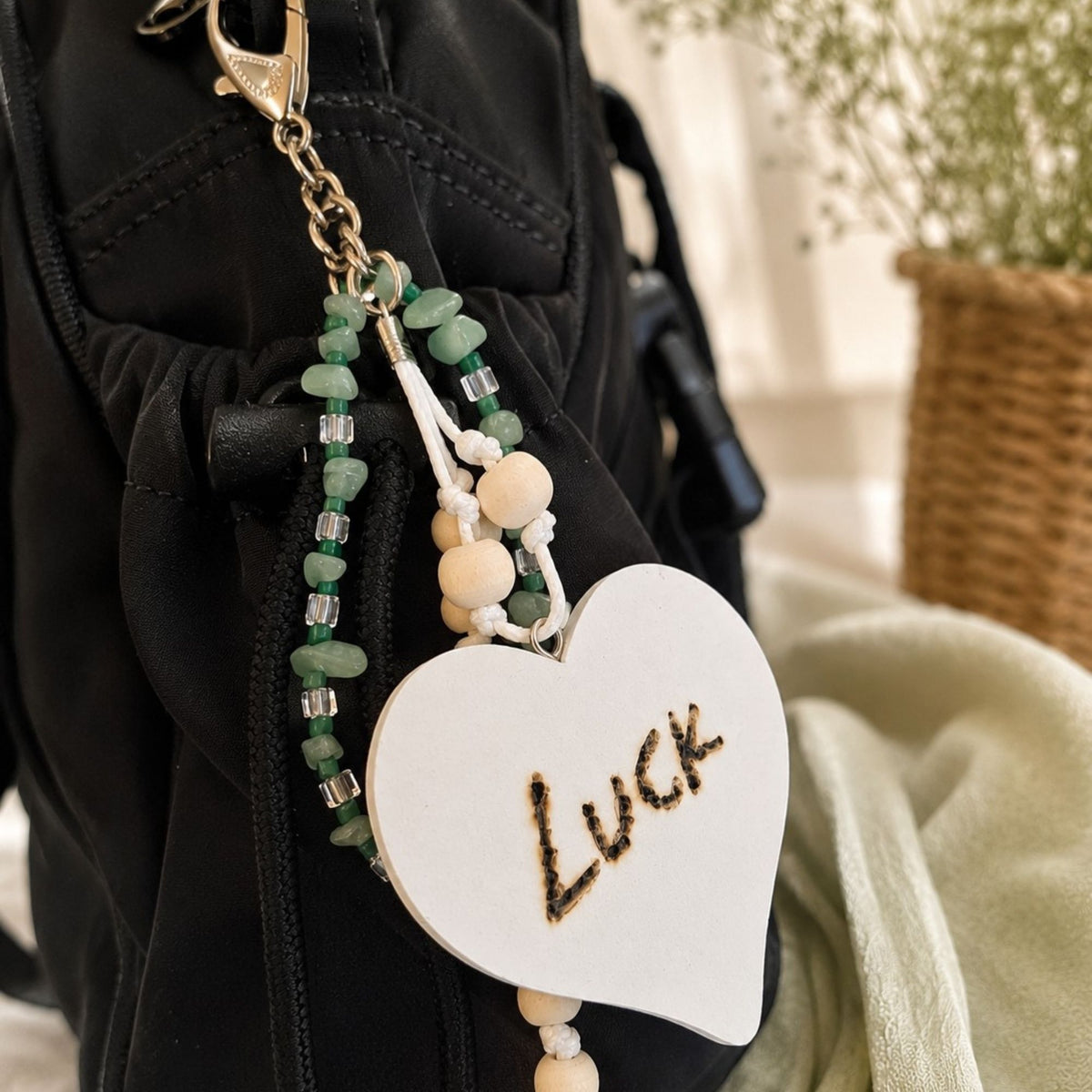 Handmade LUCK bag charm attached to a black bag, featuring green aventurine chips, dark green and clear glass beads, natural wooden beads and white cord. The large white wooden heart with the hand-burned word “LUCK” creates a striking contrast against the black fabric, finished with a silver-colored lobster clasp.