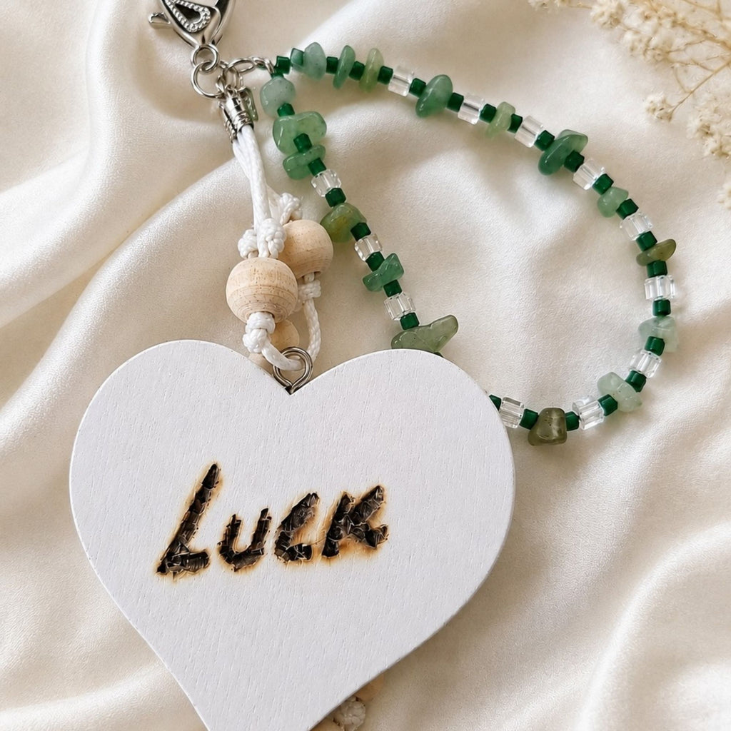 Handmade LUCK bag charm displayed on ivory satin, featuring green aventurine chips, dark green and clear glass beads, natural wooden beads and white cord. The large white wooden heart with the hand-burned word “LUCK” is shown prominently, with a silver-colored lobster clasp at the top.