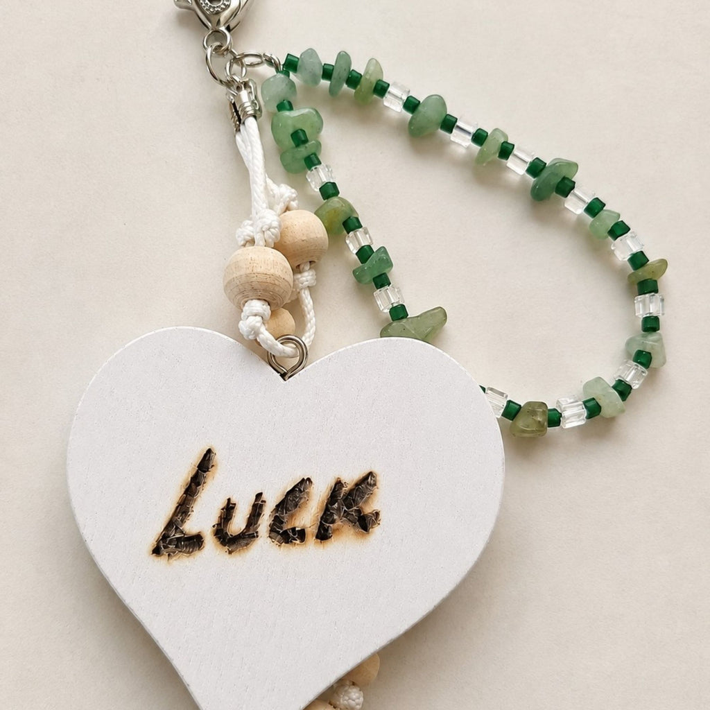 Handmade LUCK bag charm on a light neutral background, featuring green aventurine chips, dark green and clear glass beads, natural wooden beads and white cord. The large white wooden heart with the hand-burned word “LUCK” is shown in close-up, finished with a silver-colored lobster clasp.