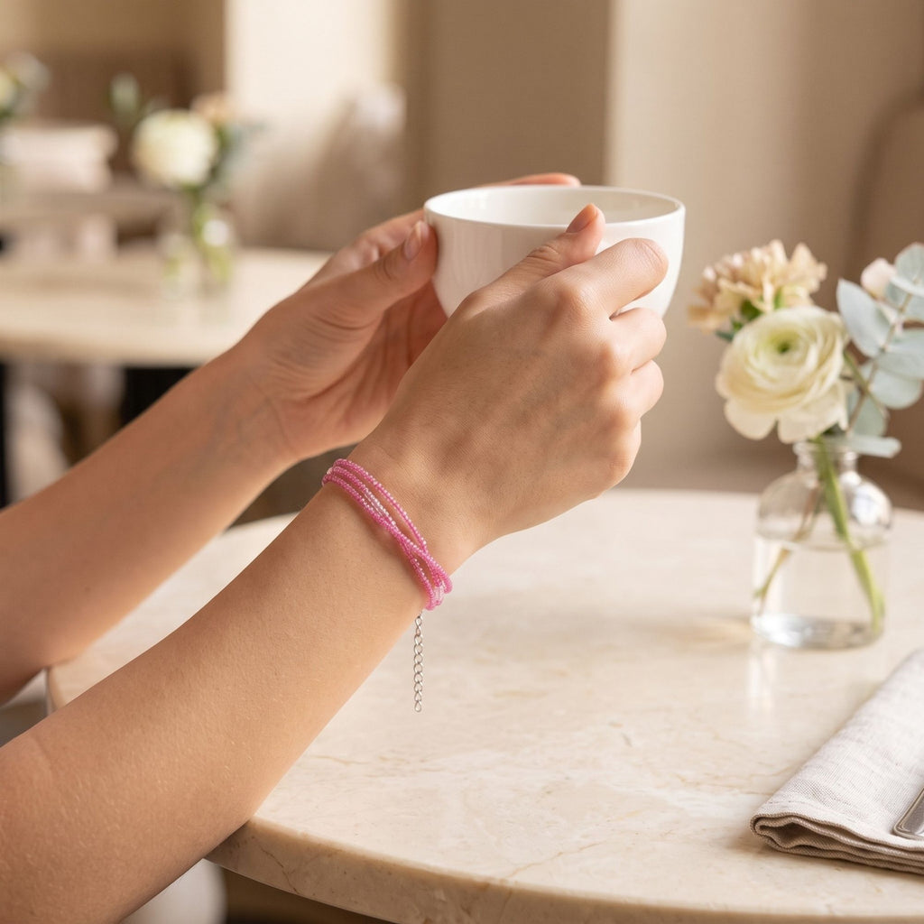 Rosalie three-strand pink glass bead bracelet worn on the wrist while holding a white cup, with a silver-tone extender chain, styled in a warm elegant café setting with soft flowers and natural light.