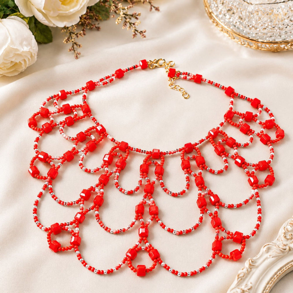 Scarlet Queen handgefertigte Statement-Halskette aus roten und transparenten Glasperlen, flach auf hellem Satin präsentiert. Das aufwendige mehrreihige Art-Déco-Design mit geschwungenen Perlenbögen sowie goldfarbenem Karabinerverschluss und Verlängerungskette ist vollständig sichtbar.