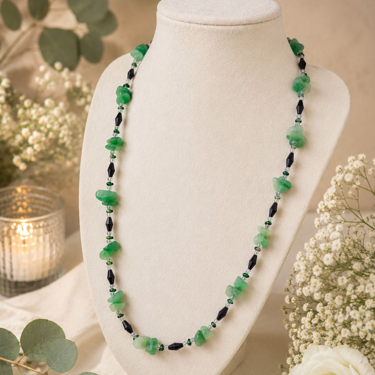 Seluna aventurine necklace displayed on a cream jewelry bust surrounded by white flowers, eucalyptus and warm candlelight. Natural green aventurine chip beads in varying shades alternate with faceted black glass beads, small dark green beads and clear accents. The full product view highlights the necklace’s delicate pattern, contrasting colors and approximately 50 cm length.