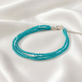 Handmade Selva three-strand bracelet featuring delicate transparent turquoise blue glass seed beads, finished with a silver-tone lobster clasp and adjustable extension chain. The vibrant turquoise beads shimmer beautifully against soft white satin, highlighting the elegant layered design and fine handcrafted details. A lightweight, minimalist bracelet inspired by clear blue water and summer days.