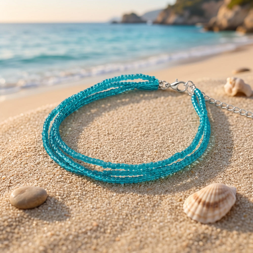 Selva handmade three-strand bracelet with delicate transparent turquoise glass seed beads, displayed on soft beach sand beside seashells. The silver-tone lobster clasp and adjustable extension chain are visible, while the turquoise beads sparkle beautifully in warm sunlight. The blue sea and sandy coastline in the background create a fresh Mediterranean-inspired setting that complements the bracelet’s elegant, lightweight summer design.