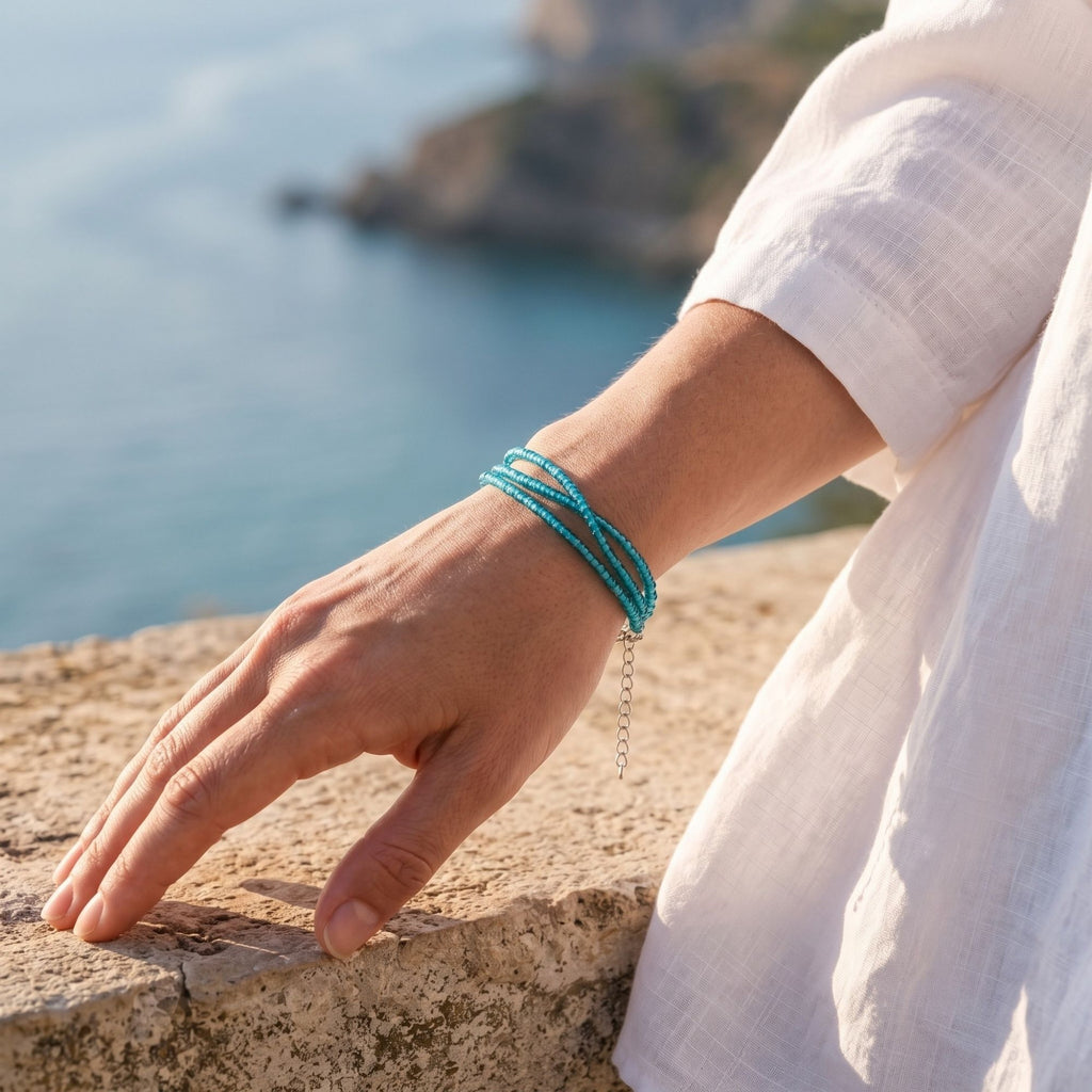 Selva handmade three-strand bracelet with delicate transparent turquoise glass seed beads, shown worn on the wrist in a sunny coastal setting. The three fine turquoise strands wrap elegantly around the wrist, while the silver-tone adjustable extension chain hangs naturally at the side. The bright blue sea and neutral linen clothing create a fresh Mediterranean summer look that highlights the bracelet’s lightweight, minimalist design.
