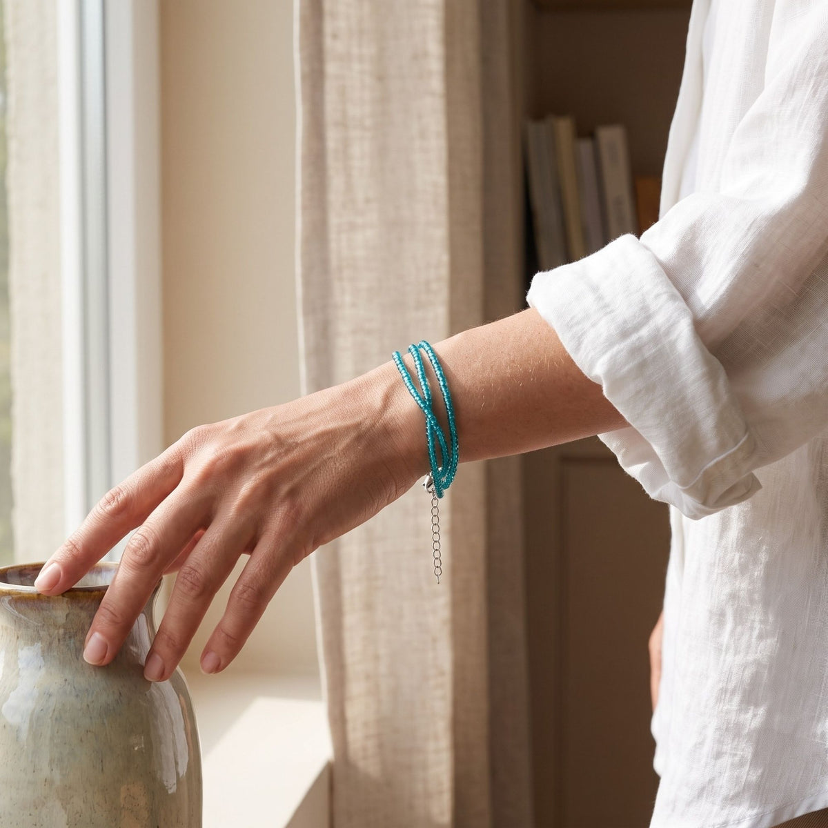Selva handmade three-strand bracelet with delicate transparent turquoise glass seed beads, worn on the wrist in a softly lit indoor setting. The three fine turquoise strands create an elegant layered look, while the silver-tone adjustable extension chain hangs naturally below the wrist. A white linen shirt and warm neutral interior emphasize the bracelet’s fresh, minimalist and versatile everyday style.