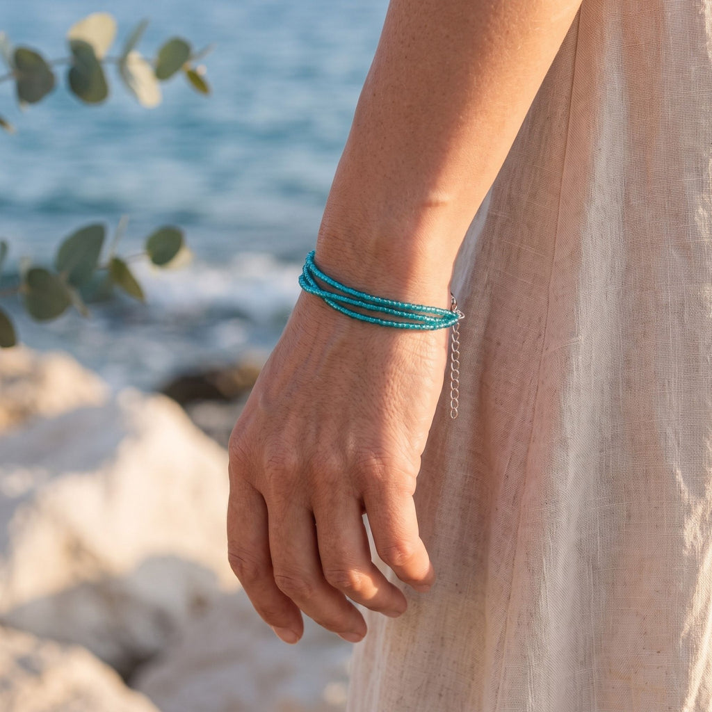 Selva handmade three-strand bracelet featuring delicate transparent turquoise glass seed beads, worn on the wrist by the sea. The three fine turquoise strands create a lightweight layered look, complemented by a silver-tone clasp and adjustable extension chain. Warm sunlight, neutral linen clothing and the soft blue ocean in the background emphasize the bracelet’s fresh, elegant and Mediterranean-inspired summer style.